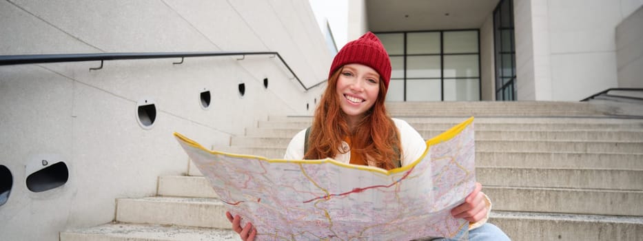 Young smiling redhead girl, tourist sits on stairs outdoors with city paper map, looking for direction, traveller backpacker explores city and looks for sightseeing.