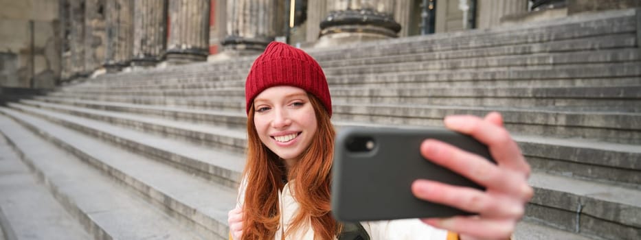 Young redhead tourist takes selfie in front of museum on stairs, holds smartphone and looks at mobile camera, makes photo of herself with phone.