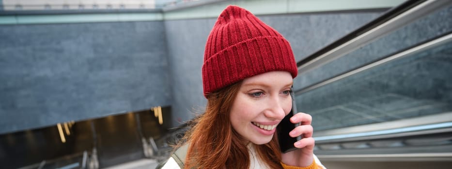 Portrait of happy redhead woman walking around town with smartphone, calling someone, talking on mobile phone outdoors.