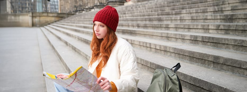 Beautiful young woman, traveler with backpack holds paper map, explores city sightseeing, plans route for tourism popular attractions.