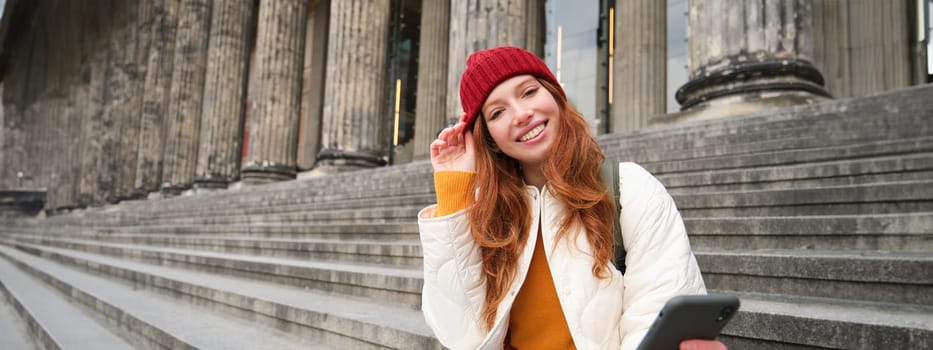 Portrait of young redhead woman in red hat, sitting on stairs, tourist looks at her mobile phone, rests on staircase of museum, connects public wifi.