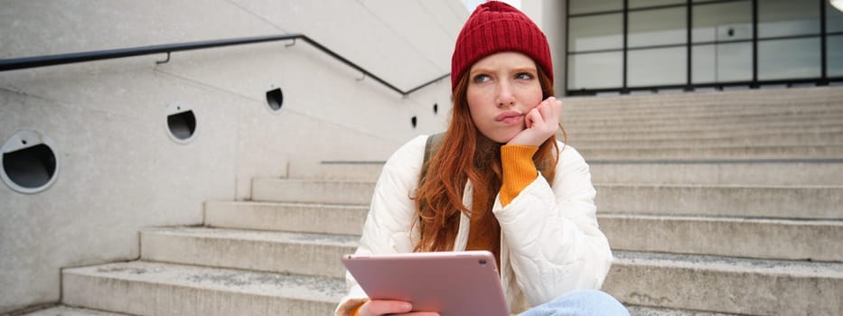 Portrait of troubled redhead girl, college student with thoughtful face, sits on street stairs, holds digital tablet, thinks how to reply on message.