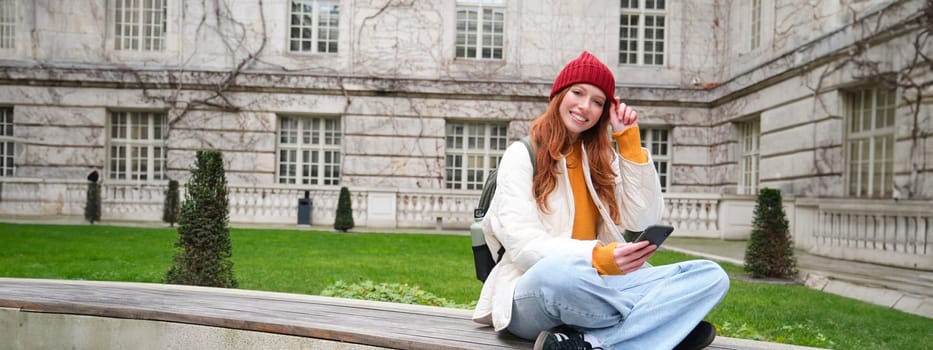 Young smiling redhead girl sits on bench and uses smartphone app, reads news online, watches video on mobile phone while relaxed in park.