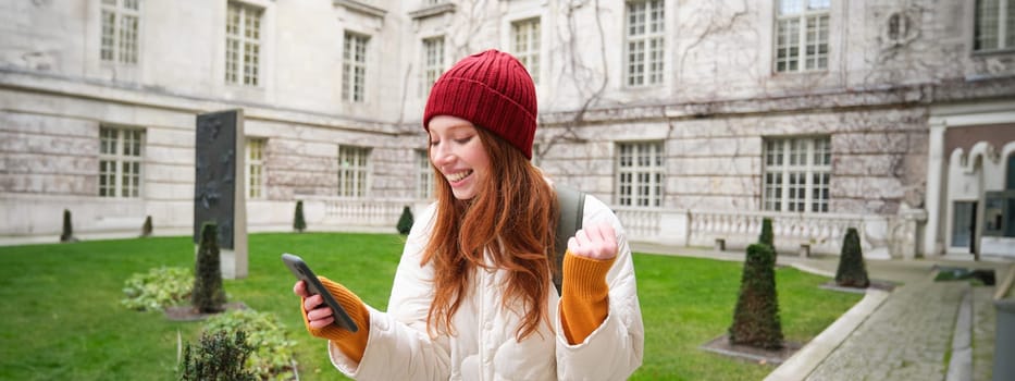 Portrait of cheerful redhead woman with smartphone, celebrating, reading amazing news and rejoicing, triumphing on street.