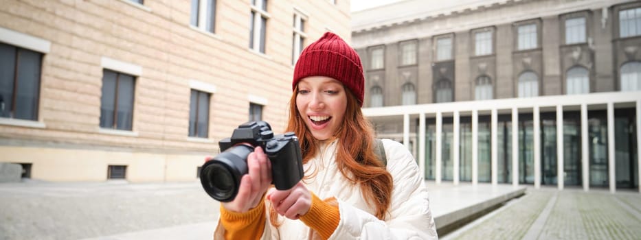 Beautiful readhead girl, photographer with professional camera takes pictures outdoors, walking around city and taking photos, sightseeing.