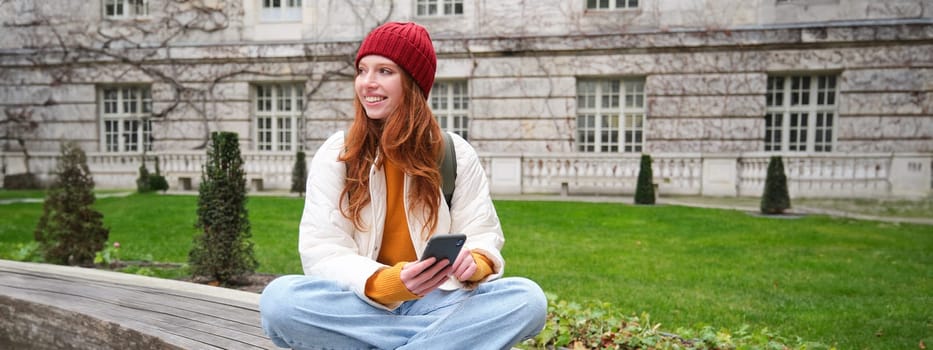 Young smiling redhead girl sits on bench and uses smartphone app, reads news online, watches video on mobile phone while relaxed in park.