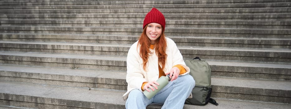 Smiling tourist, girl sits on stairs, rests on staircase, takes thermos from backpack, drinks hot coffee from flask.