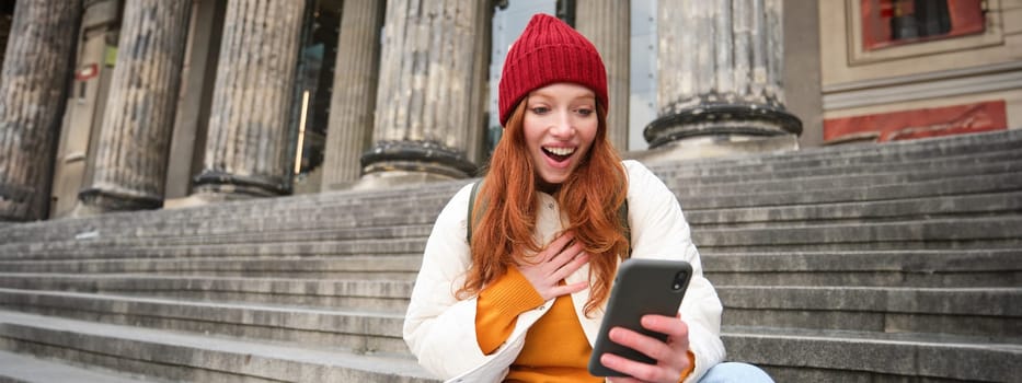Redhead female tourist talks on mobile phone, video chatting with friends, says hi and waves hand at smartphone camera.