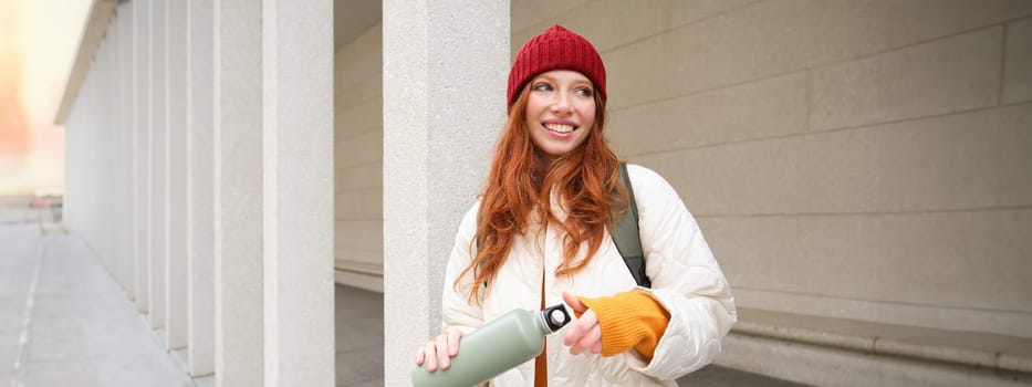 Young happy redhead woman in red hat, drinking from thermos, warming up with hot drink in her flask while walking around city, tourist relaxes with warm refreshemtn.