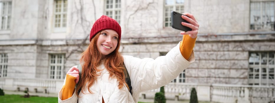 Cute ginger girl in red hat takes selfie during her tourist trip abroad. Young redhead woman makes a photo of herself in front of historical landmark.