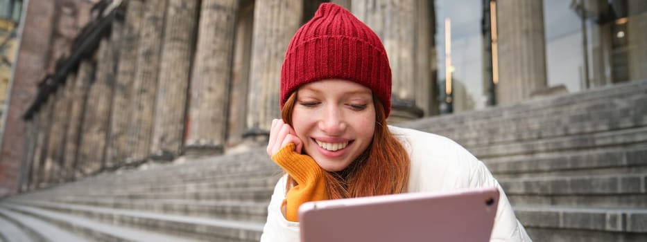 Portrait of young redhead woman sitting outdoors on stairs, reading e-book on digital tablet, wearing red hat and warm clothes.