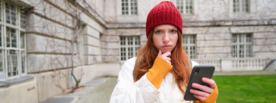 Portrait of young redhead woman in red hat, holds smartphone and looks thoughtful, frowning with perplexed, thinking face expression, standing on street near building.
