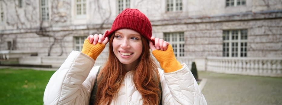 Portrait of smiling redhead woman puts on red hat and smiles, wearing warm clothes while exploring city streets.