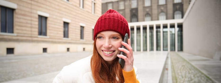 Stylish modern redhead girl talks on mobile phone, makes a telephone call, calling someone on smartphone app from outside, stands on street and smiles.