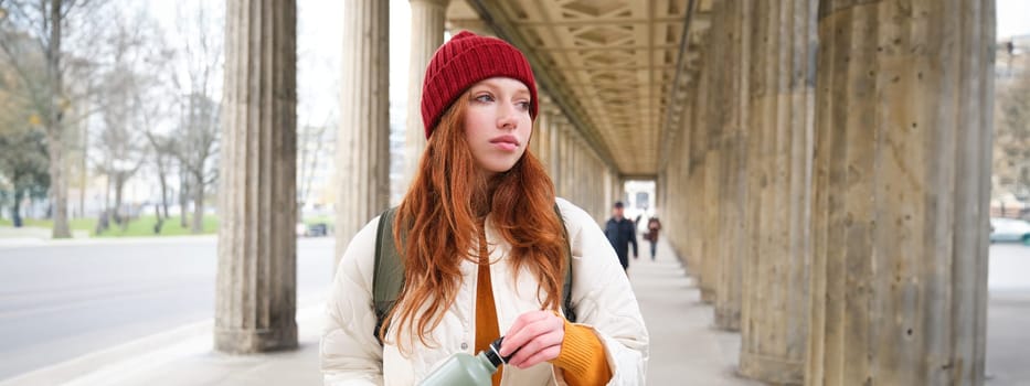 Young redhead girl holds thermos in hands, pours herself a hot drink while walking in city. Tourist takes break, opens flask for refreshment beverage.