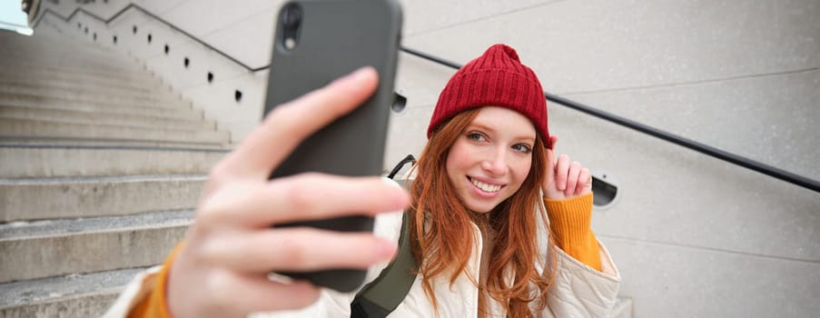 Mobile phone and people lifestyle. Stylish redhead girl takes selfie on her smartphone, poses for photo with mobile phone in hand, smiles happily, sits on stairs outdoors.