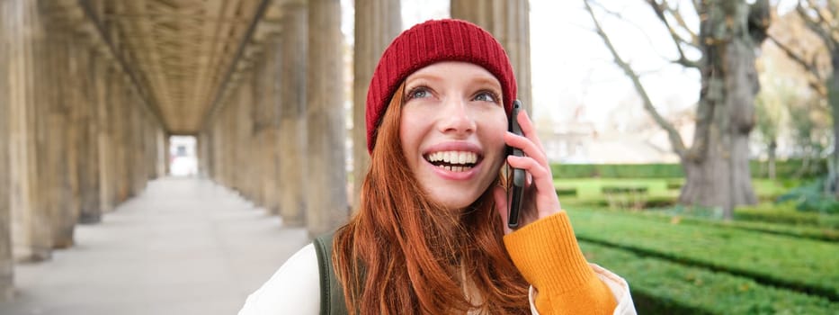 Portrait of redhead european girl in red hat, makes a phone call, walks in city and talks to friend on smartphone. Copy space