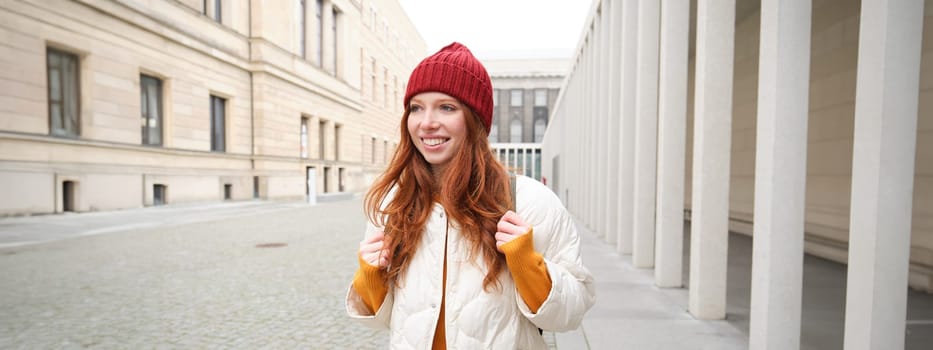 Female tourist in red hat with backpack, sightseeing, explores historical landmarks on her trip around europe, smiling and posing on street.