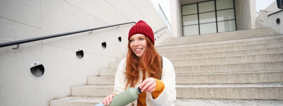 Smiling traveler, redhead girl tourist sits on stairs with flask, drinks hot coffee from thermos while travelling and sightseeing around foreign city, sits on stairs and rests.