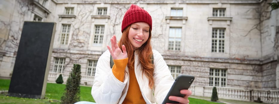 Smiling young woman says hello on video chat, connects public wifi in park and talks someone online with smartphone app.