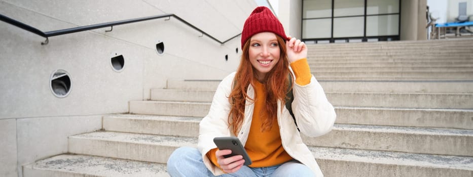 Beautiful redhead girl, student sits with her mobile phone on stairs near campus, smiles, wears red hat, uses smartphone city app.