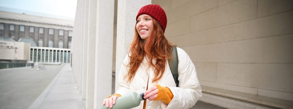 Young happy redhead woman in red hat, drinking from thermos, warming up with hot drink in her flask while walking around city, tourist relaxes with warm refreshemtn.