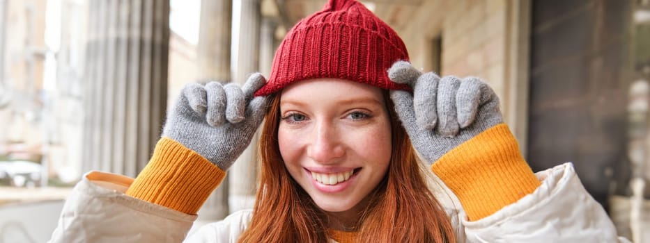 Headshot of happy redhead girl with freckles, wears red hat and gloves in winter, walks around city on chilly weather and smiles.