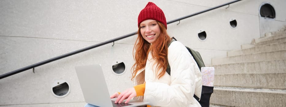 Portrait of young girl traveller, sitting with backpack and map of city, working on laptop, connecting to public wifi and sitting on stairs outdoors, using internet to book hotel room.