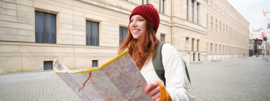 Beautiful redhead woman, tourist with city map, explores sightseeing historical landmark, walking around old town, smiling happily.