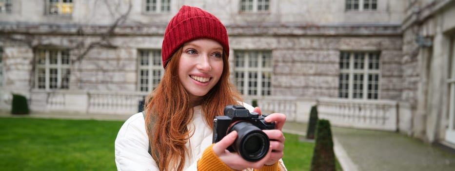Happy redhead girl tourist, takes photos, photographer with professional camera walks around city and captures beautiful pictures.