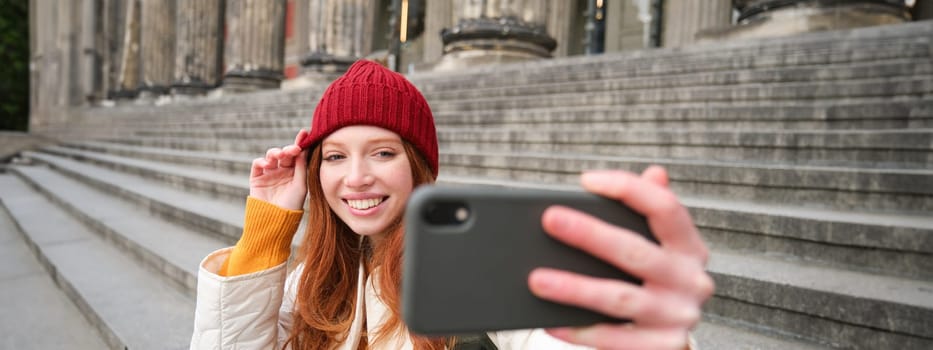 Young redhead tourist takes selfie in front of museum on stairs, holds smartphone and looks at mobile camera, makes photo of herself with phone.