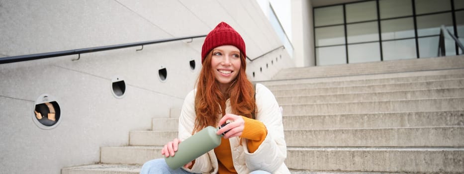 Smiling traveler, redhead girl tourist sits on stairs with flask, drinks hot coffee from thermos while travelling and sightseeing around foreign city, sits on stairs and rests.