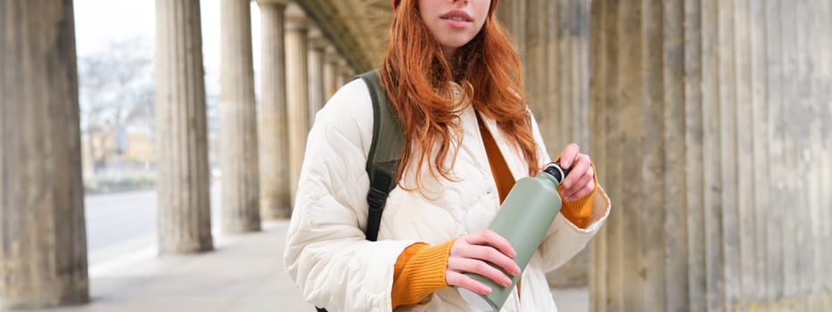 Close up portrait of redhead girl, tourist stands on street, opens thermos with hot drink, rests and pours herself coffee from flask.