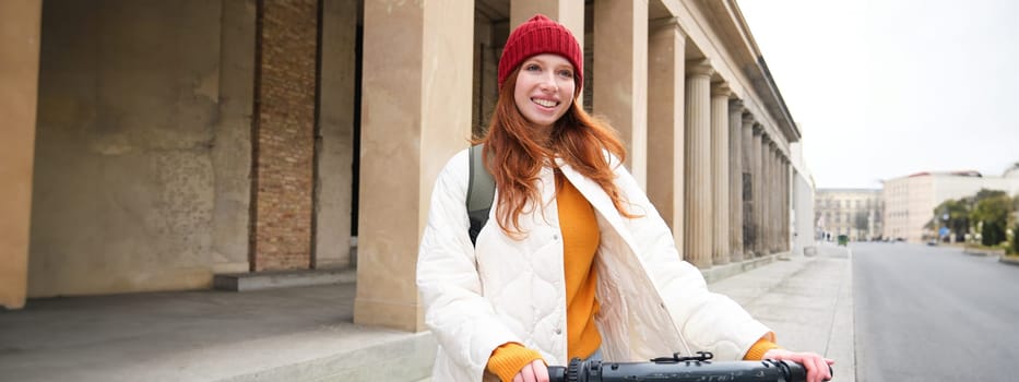 Smiling redhead european girl drives public e-scooter, tourist explores city, rides in city centre.