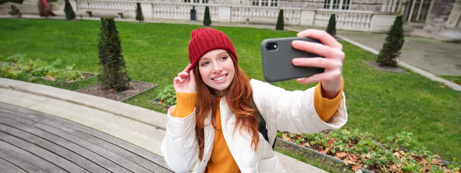 Young teen redhead girl sits on bench in park and takes selfie, makes a photo of herself with smartphone app, records vlog.