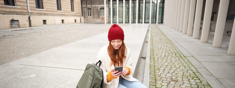 Young redhead woman with smartphone, sitting outdoors with backpack, student looking at her mobile phone, texts message.