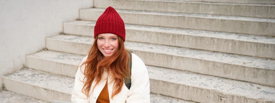 Smiling redhead girl, student sits on stairs outdoors and uses laptop, connects to public wifi in city and works on project, uses internet on computer.