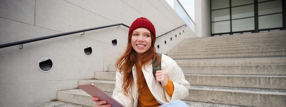 Young beautiful girl with long red hair, smiles, uses internet application, connect to wifi near campus, sits on stairs with digital tablet.