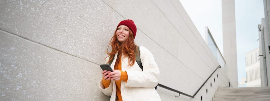 Happy girl student in red hat, holds smartphone, tourist looks at map app on her phone, explores sightseeing, texts message, looks for couchsurfing, rents place to stay online.