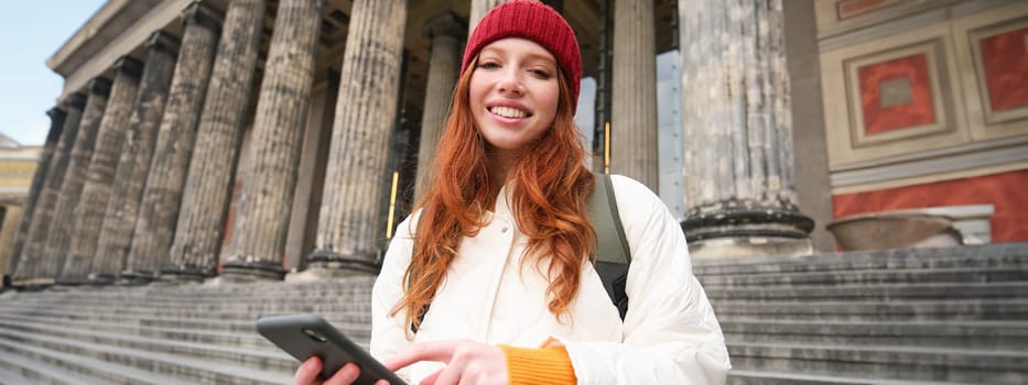 Portrait of young urban girl with backpack, using smartphone, walking around city, looking at map on mobile app, checking direction.