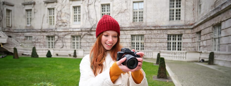 Happy redhead girl tourist, takes photos, photographer with professional camera walks around city and captures beautiful pictures.
