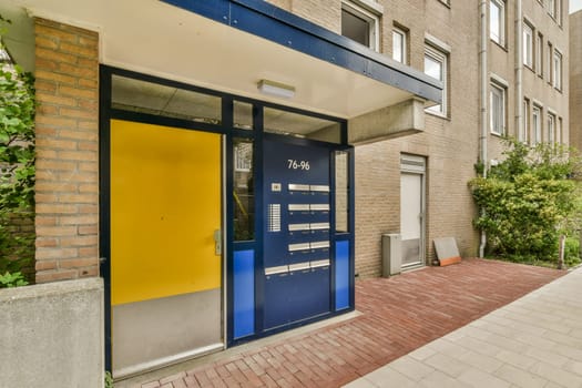 a blue and yellow door on the side of a brick building in an urban area with trees, bushes and plants