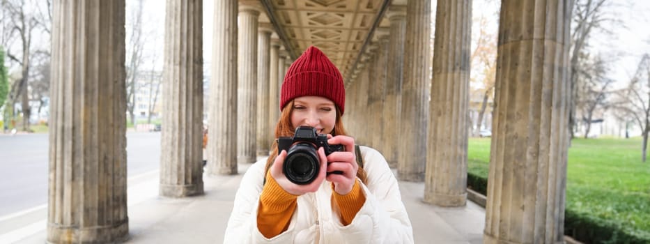 Young redhead female photographer, makes lifestyle shooting in city centre, takes photos and smiles, looks for perfect shot, makes picture.