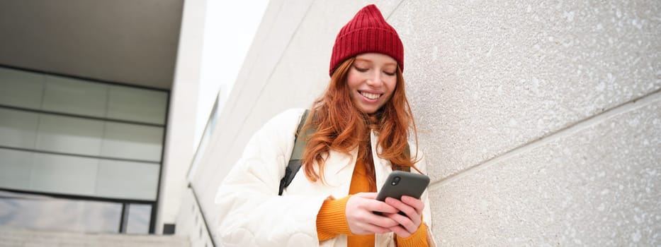 Happy girl student in red hat, holds smartphone, tourist looks at map app on her phone, explores sightseeing, texts message, looks for couchsurfing, rents place to stay online.