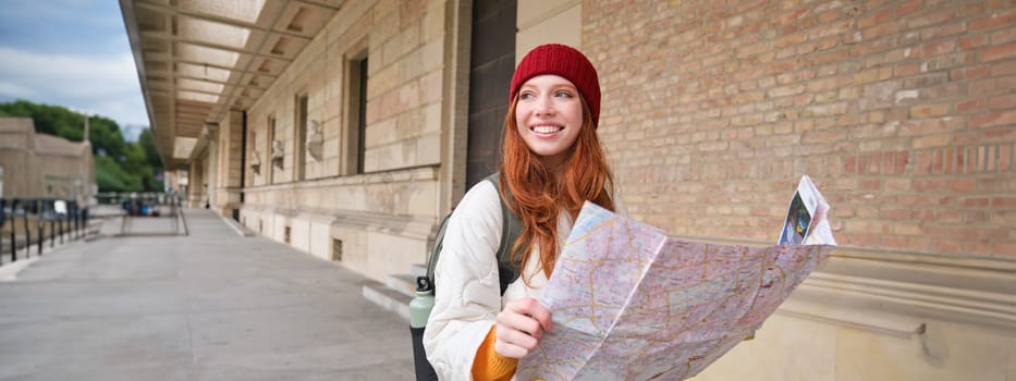 Adventurous redhead girl walks in town with paper map, explores city as tourist, looks for popular tourism attractions, looks around excited and smiles.