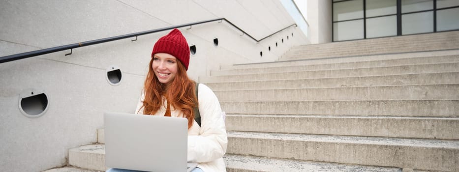 Smiling redhead girl, student sits on stairs outdoors and uses laptop, connects to public wifi in city and works on project, uses internet on computer.