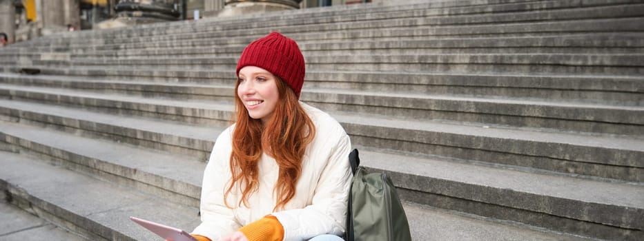 Outdoor shot of young stylish redhead girl sits on staircase and connects to public wifi, uses digital tablet, reads news on gadget.