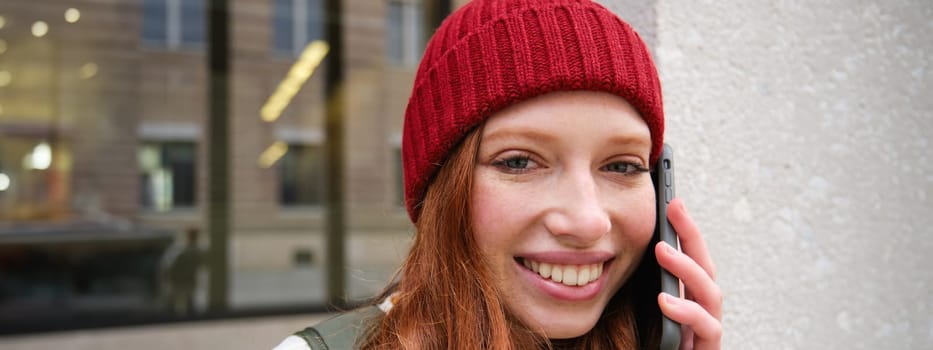 Young people and mobile connection. Happy redhead girl talks on phone, makes telephone call, stands outdoors with backpack and uses smartphone app.