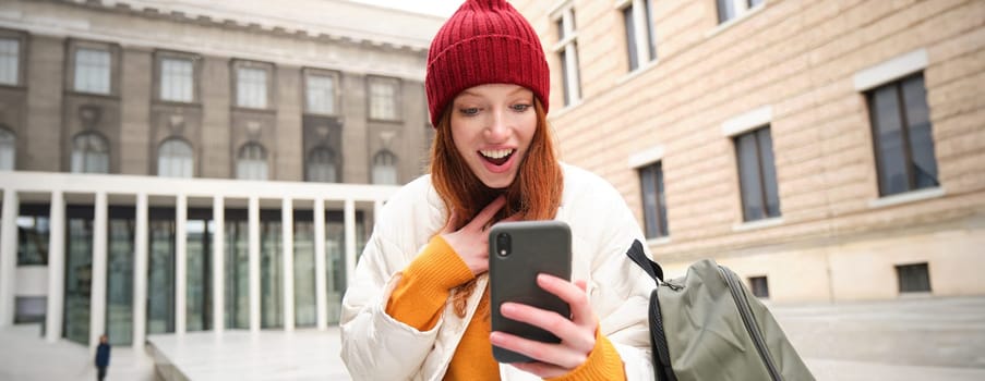 Young redhead woman with smartphone, sitting outdoors with backpack, student looking at her mobile phone, texts message.