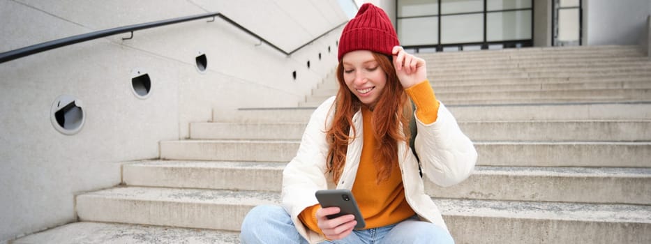 Stylish european girl with red hair, sits on public stairs with smartphone, places online order, sends message on mobile phone social app, smiles happily.